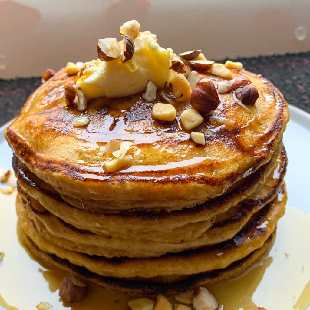 A stack of pumpkin pancakes, topped with maple syrup, butter and hazelnuts.