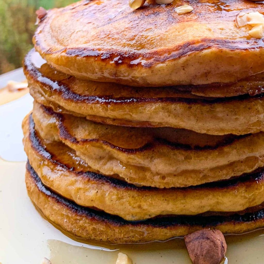 A close-up of a stack of pumpkin pancakes, topped with maple syrup, butter and hazelnuts. 