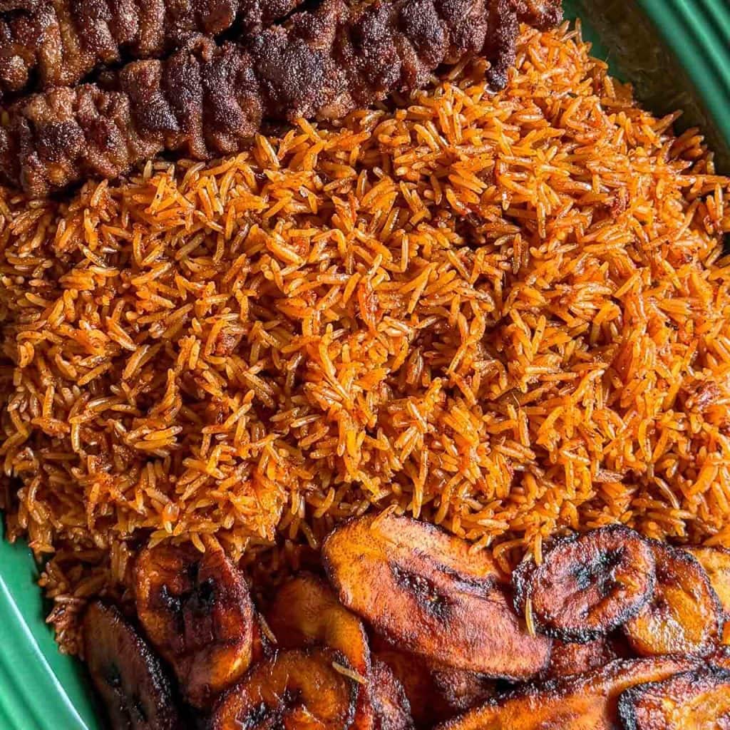 A closeup shot of mum's Jollof Rice, served with fried plantain and beef suya on a green platter.