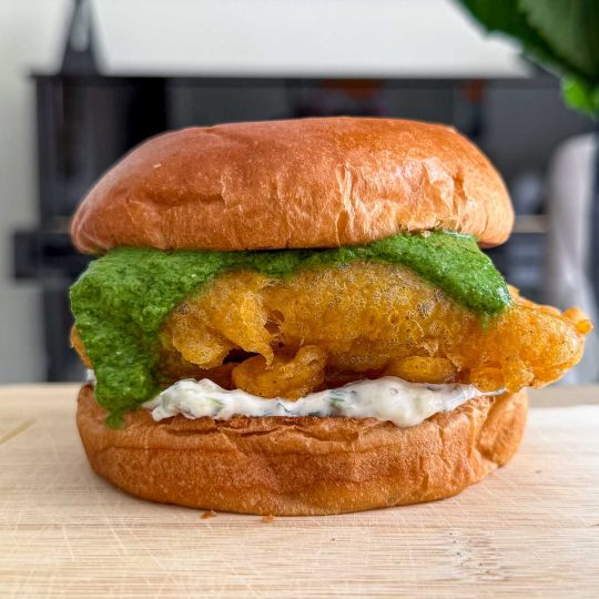 Golden curried fish fillet tucked into a brioche bun with cucumber raita and coriander chutney spilling over the sides.