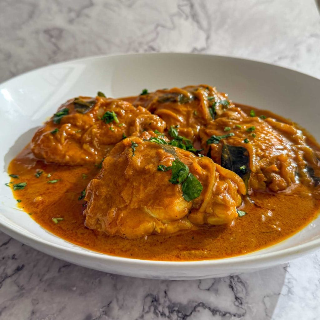 A side-on view of Caramelised Onion Chicken Curry served in a white shallow bowl, with golden chicken thighs nestled in a rich, onion-forward coconut sauce.