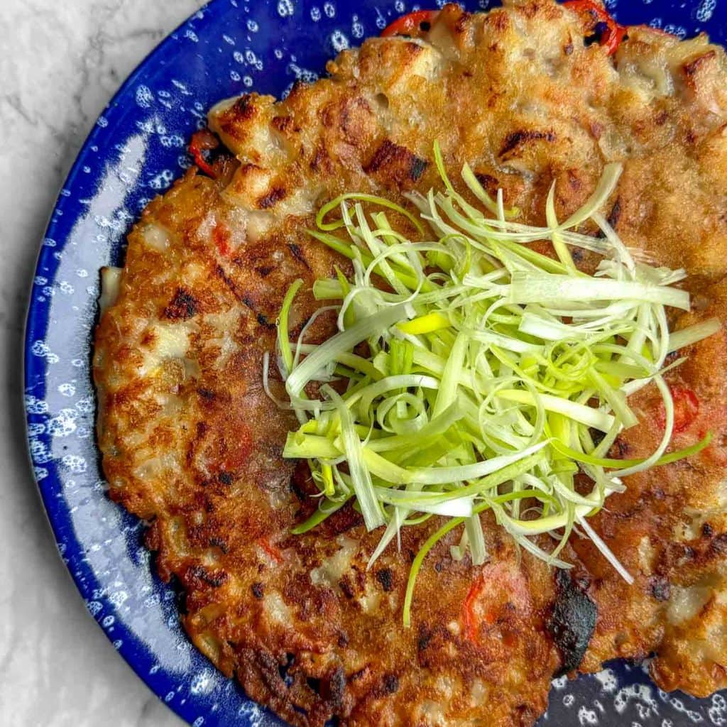 A closeup of Korean chicken pancake served on a blue plate speckled with white, topped with a mound of fresh spring onions in the centre