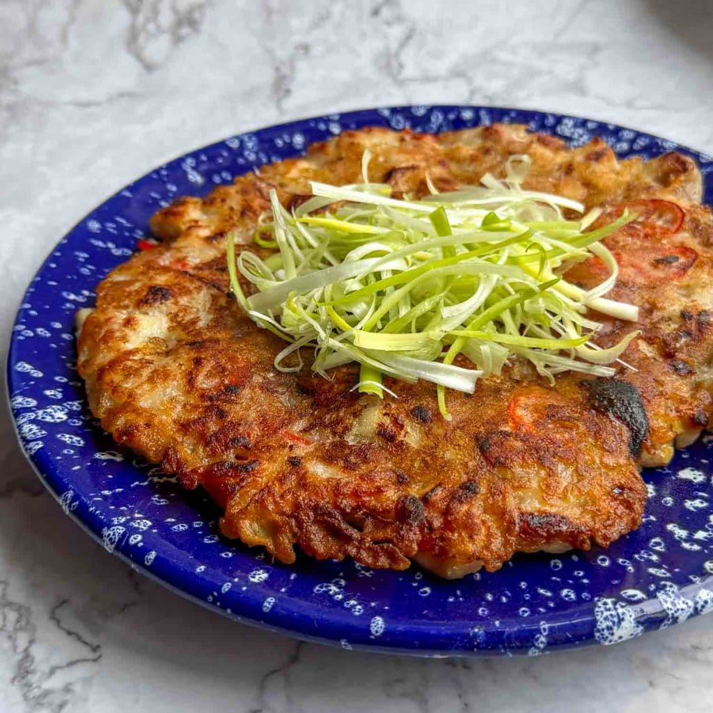 Golden, crispy Korean chicken pancake on a blue plate speckled with white, showing crisp edges and topped with soaked spring onions.