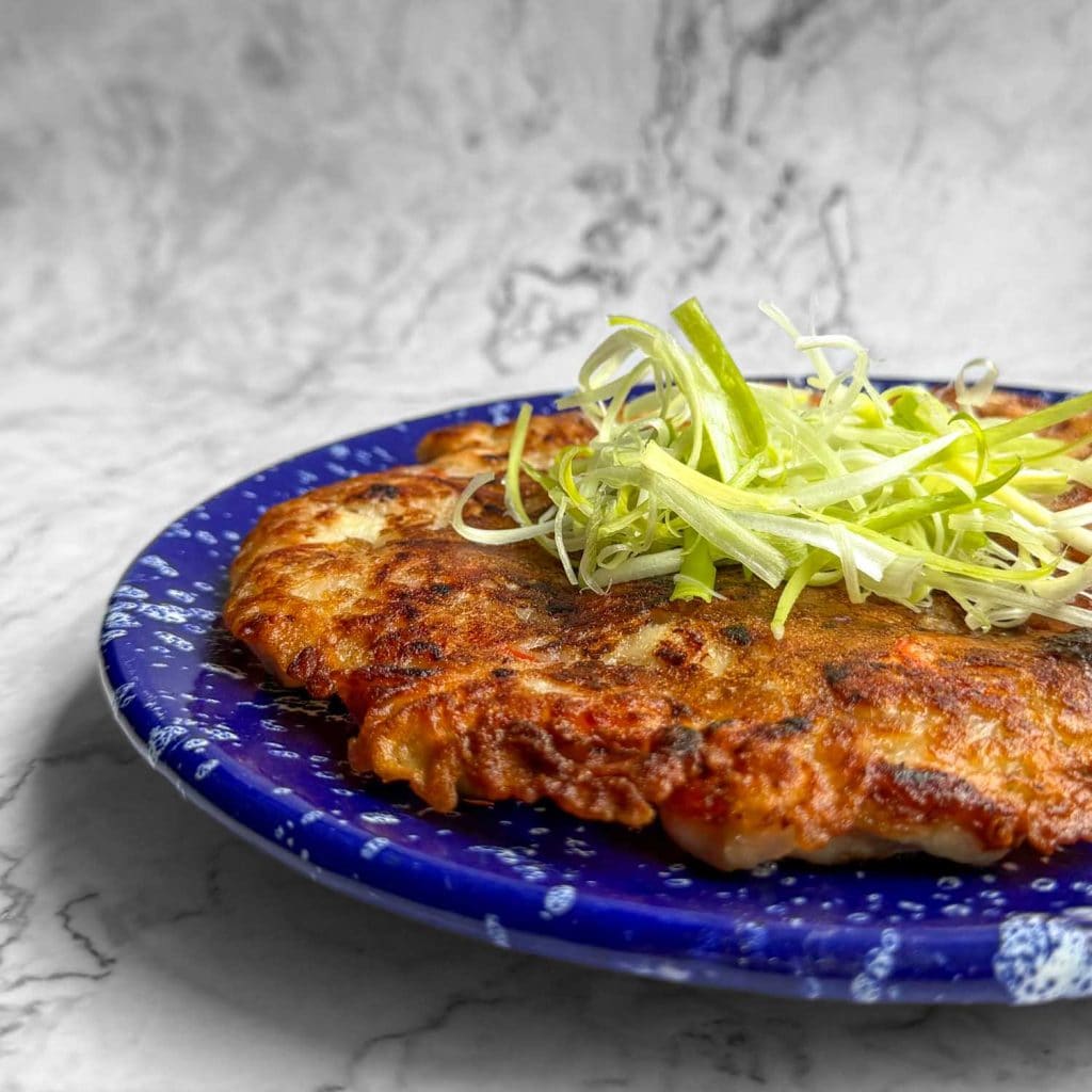 Golden Korean chicken pancake on a blue plate speckled with white, showing crisp edges and topped with spring onions.