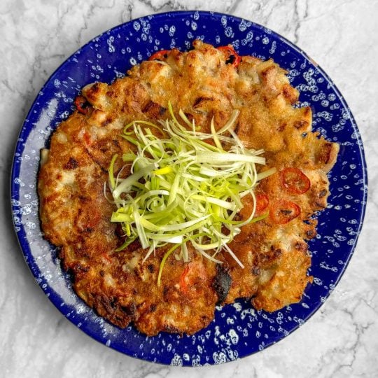 A Korean chicken pancake served on a blue plate speckled with white, topped with a mound of fresh spring onions in the centre.