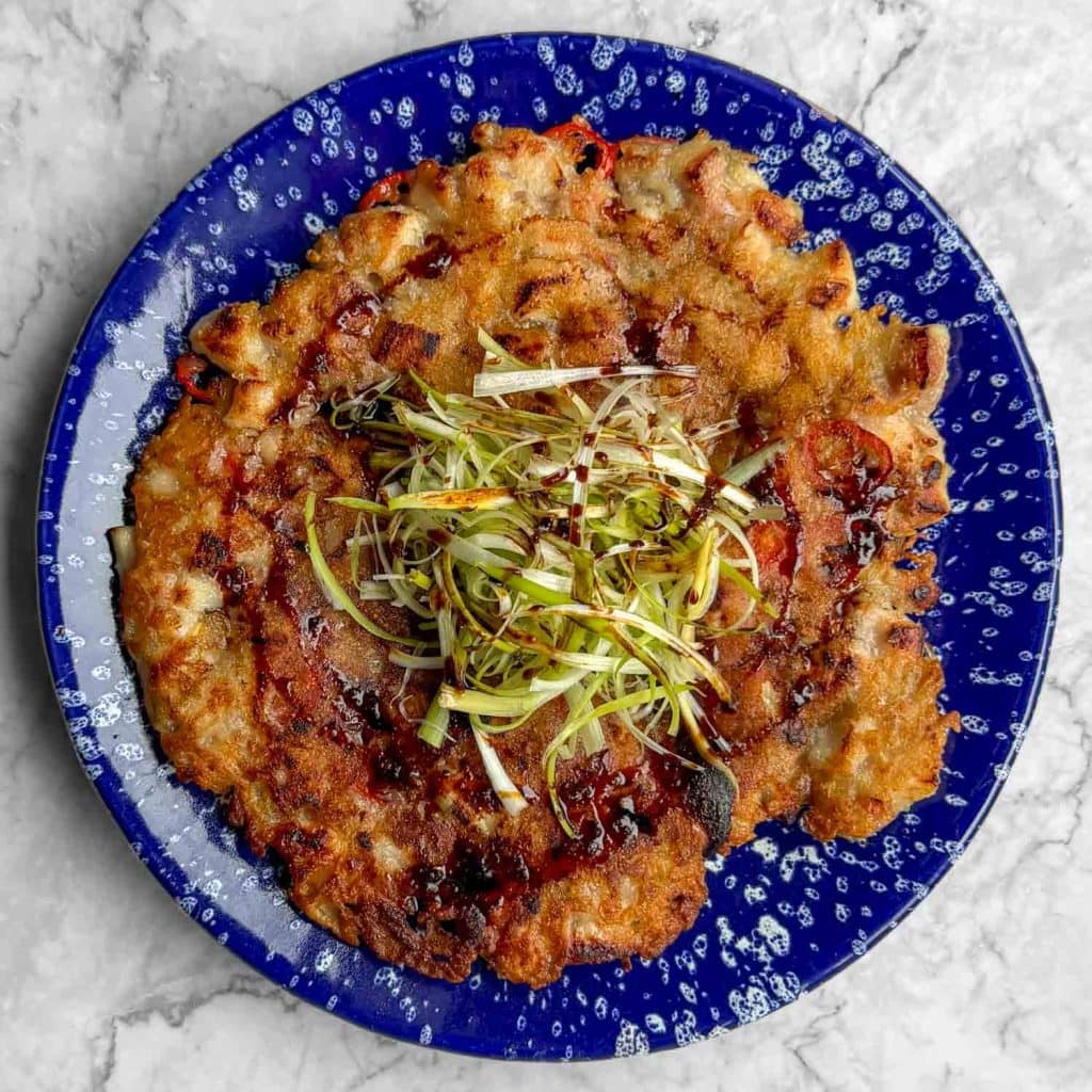 Korean chicken pancake on a blue speckled plate, topped with spring onions and drizzled with a soy and chilli dipping sauce.