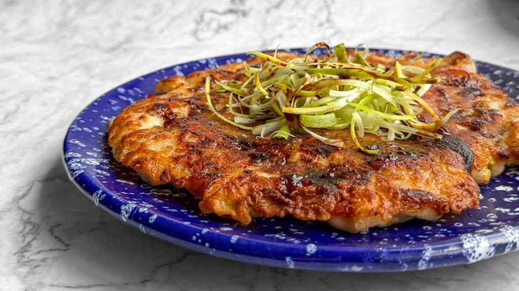 Crispy Korean chicken pancake topped with spring onions and a glossy soy-based sauce, served on a blue plate speckled with white.