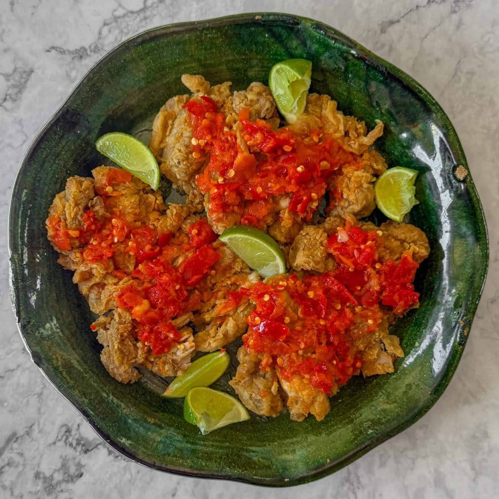 Ayam geprek, aka Indonesian smashed fried chicken, topped with sambal and served on a green platter with lime wedges.