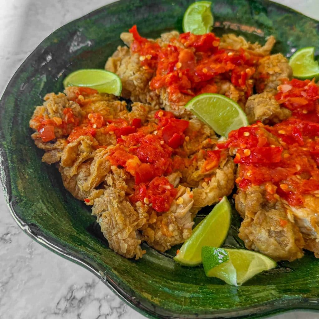 A close up of ayam geprek, aka Indonesian smashed fried chicken, topped with sambal and served on a green platter.