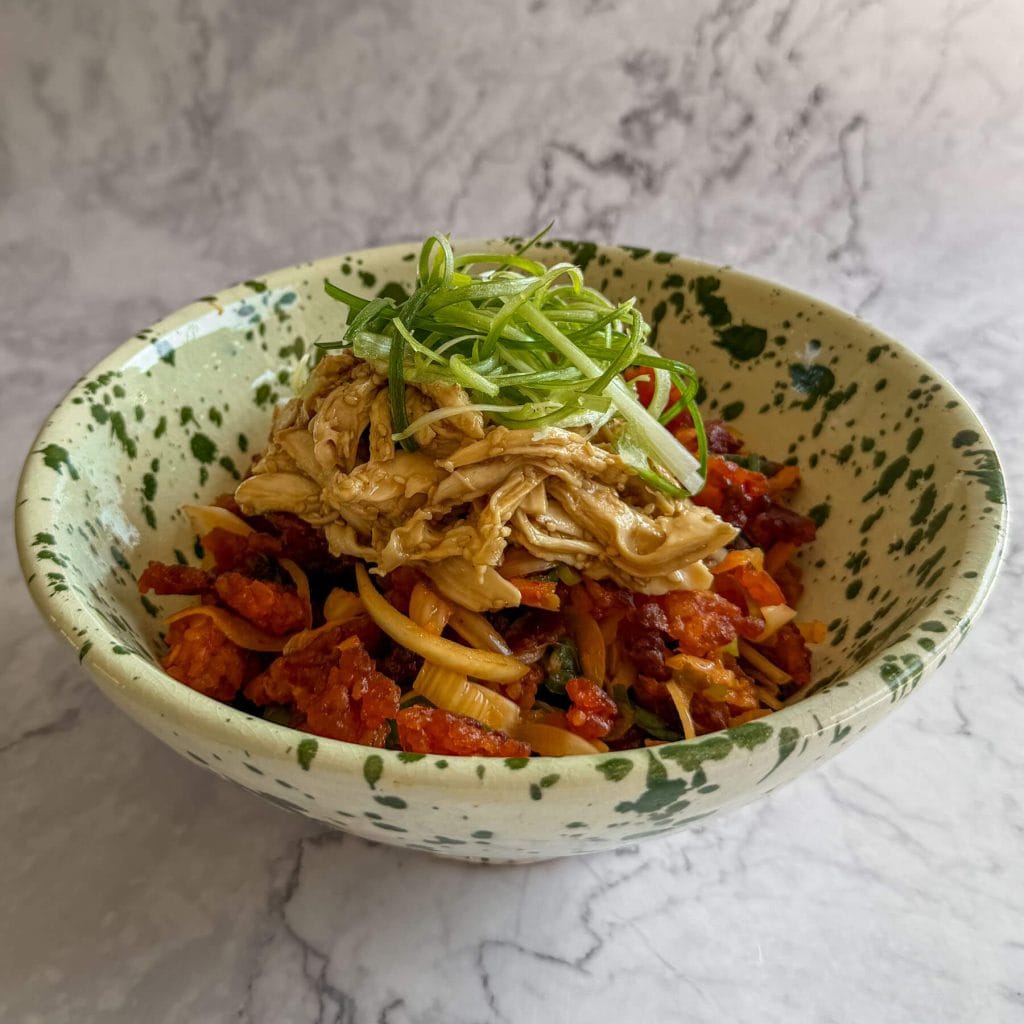 Gochujang crispy rice, topped with pulled sesame chicken and sliced spring onions, served in a white speckled bowl.